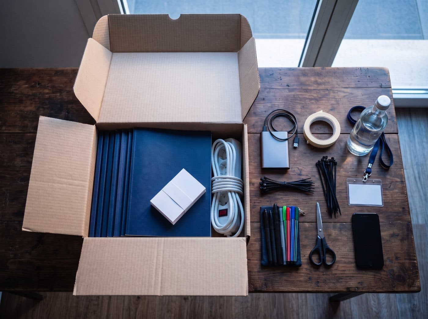 An open box with brochures, business cards and a power strip for a trade show booth, next to tape, cable ties, markers, a badge and a phone